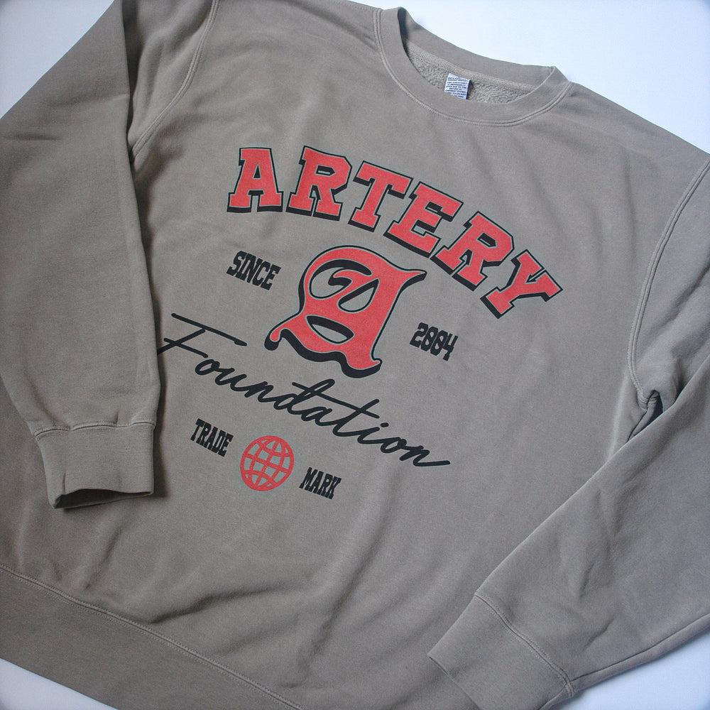 A classic-fit beige crewneck sweatshirt by Artery Foundation, featuring red and black ARTERY Foundation Since 2004 text, a bold central "A," TRADE MARK, a red globe icon, and made from soft ring-spun cotton for comfort.
