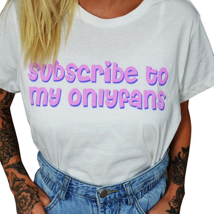 A Stay Sick Clothing white cropped shirt with bold pastel purple and pink Subscribe to my OnlyFans text, paired with high-waisted blue jeans. Tattoos are visible on the wearer’s arms.