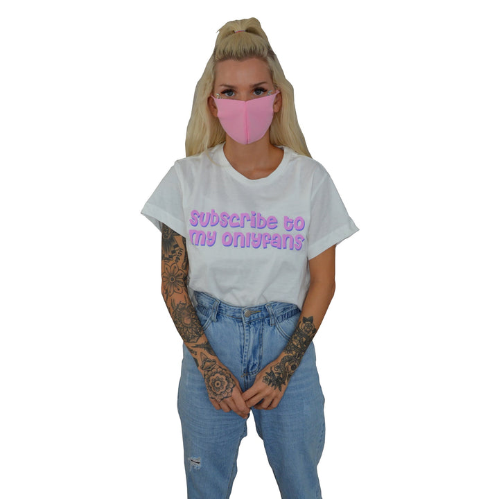 A blonde woman wearing a pink face mask and Stay Sick Clothing’s white cropped shirt with Subscribe to my onlyfans in purple stands with her hands together against a white background.