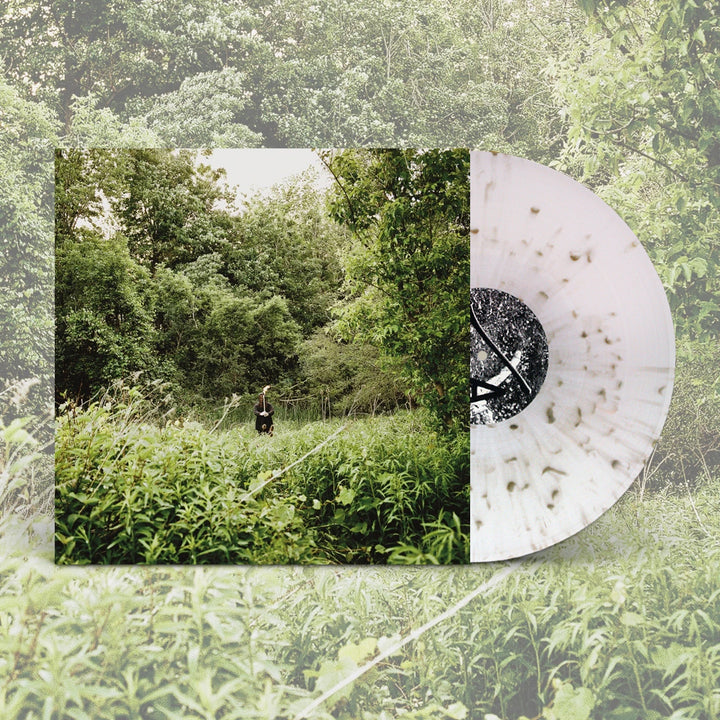 A white vinyl with dark streaks by Exalt sits partly in the Flesh To Ash album cover, which shows a person standing in tall green grass amid dense trees. The background displays a faded, enlarged version of this cover image.