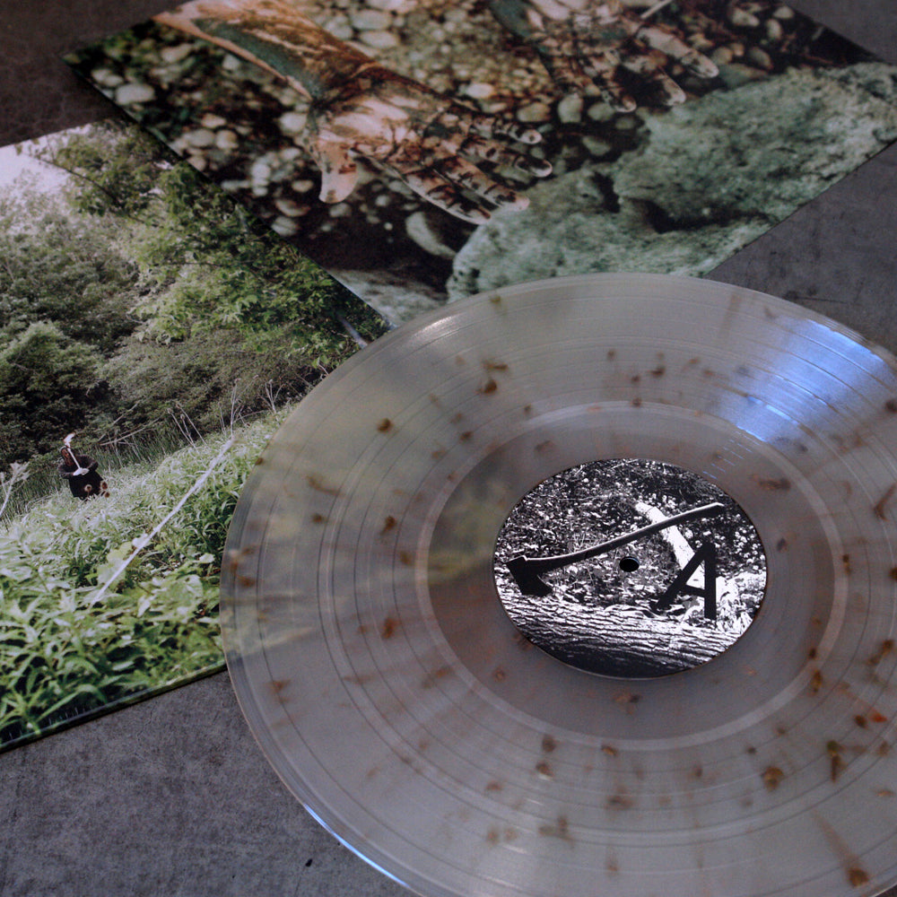 A clear Exalt Palmcarver vinyl record with brown speckles rests on a gray surface, partly covering its "Death Is A Road" album cover, which displays nature-themed photos of greenery, trees, and a person in tall grass.
