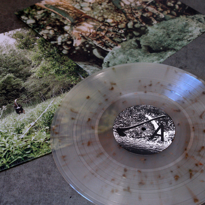 A clear Exalt Palmcarver vinyl record with brown speckles rests on a gray surface, partly covering its "Death Is A Road" album cover, which displays nature-themed photos of greenery, trees, and a person in tall grass.