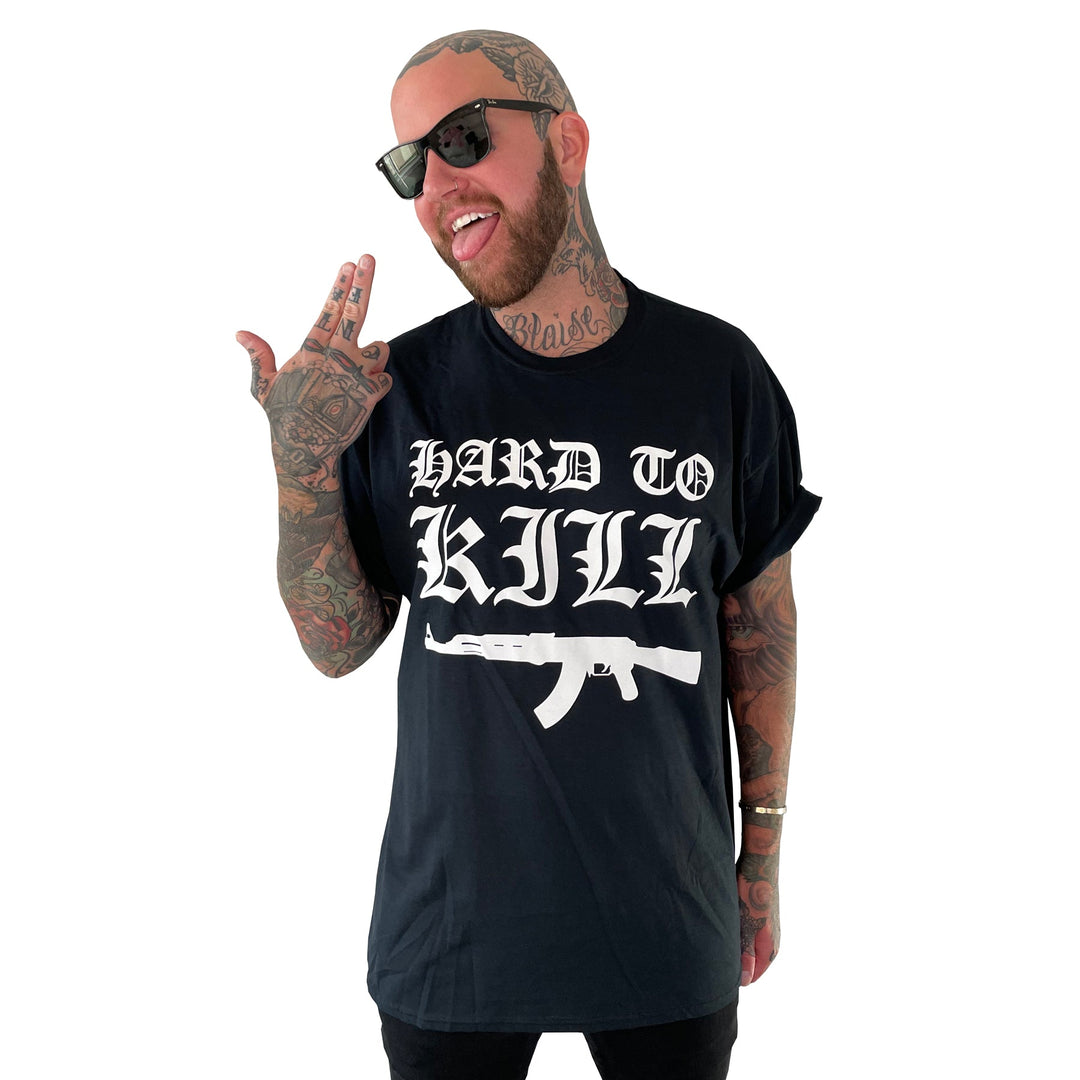 A tattooed man in sunglasses and a black HARD TO KILL t-shirt with a rifle illustration from Stay Sick Clothing smiles, making a hand gesture near his head against a white background.