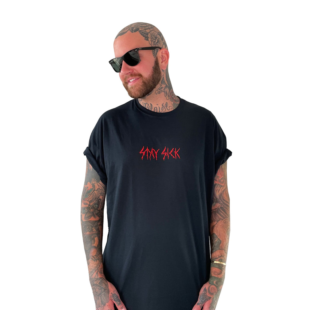A tattooed person in sunglasses and a black Stay Sick Clothing T-shirt with red STAY SICK text stands smiling against a white background, looking to the side.