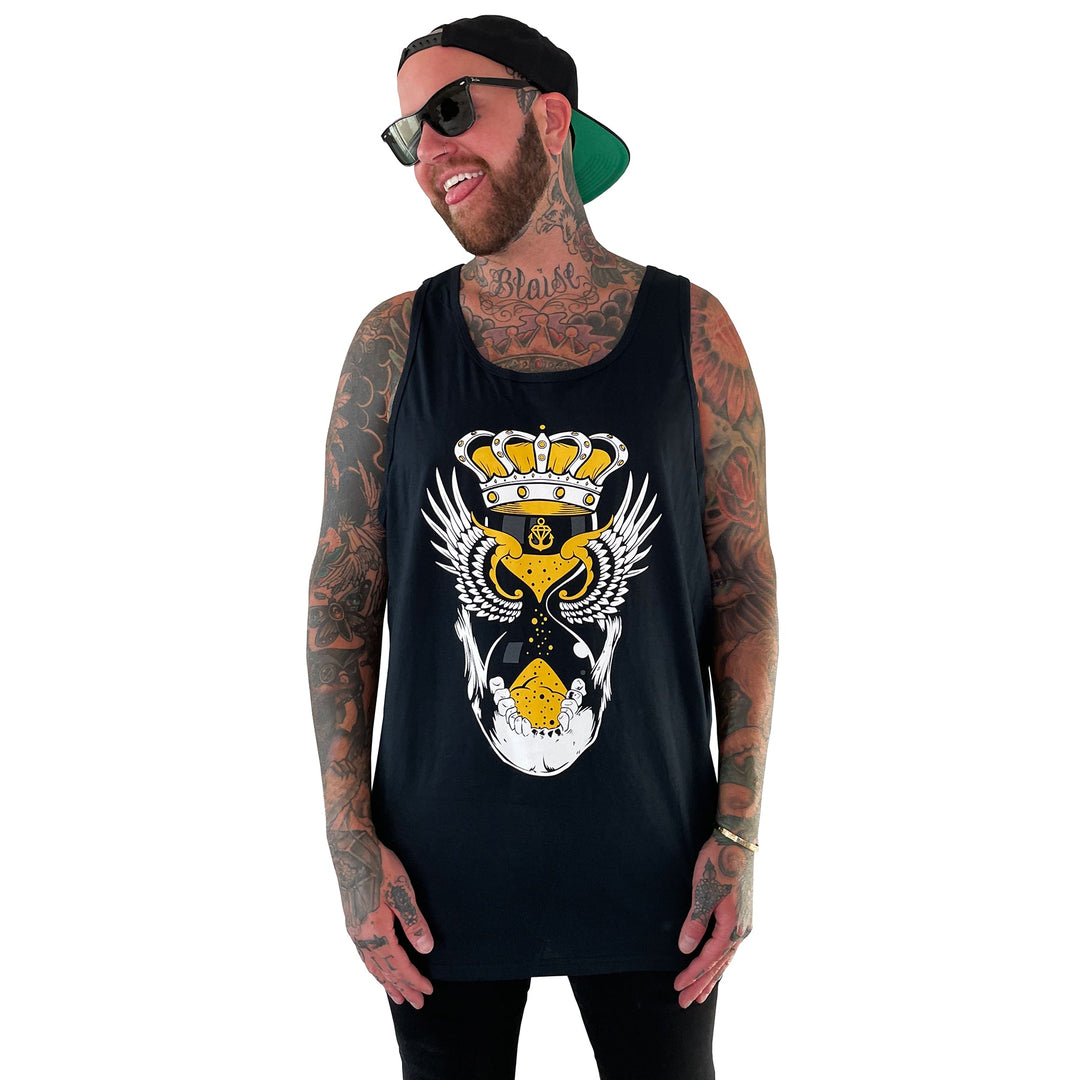 A smiling tattooed man in sunglasses, a backward cap, and a black tank top featuring a crowned heart with wings stands against a white background. The tank top is by Stay Sick Clothing.