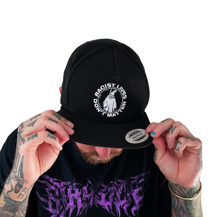 A person with hand tattoos adjusts a Stay Sick Clothing black snapback hat that says Racist Lives Dont Matter in white, with a central illustration. They wear a black shirt with purple lettering against a white background.