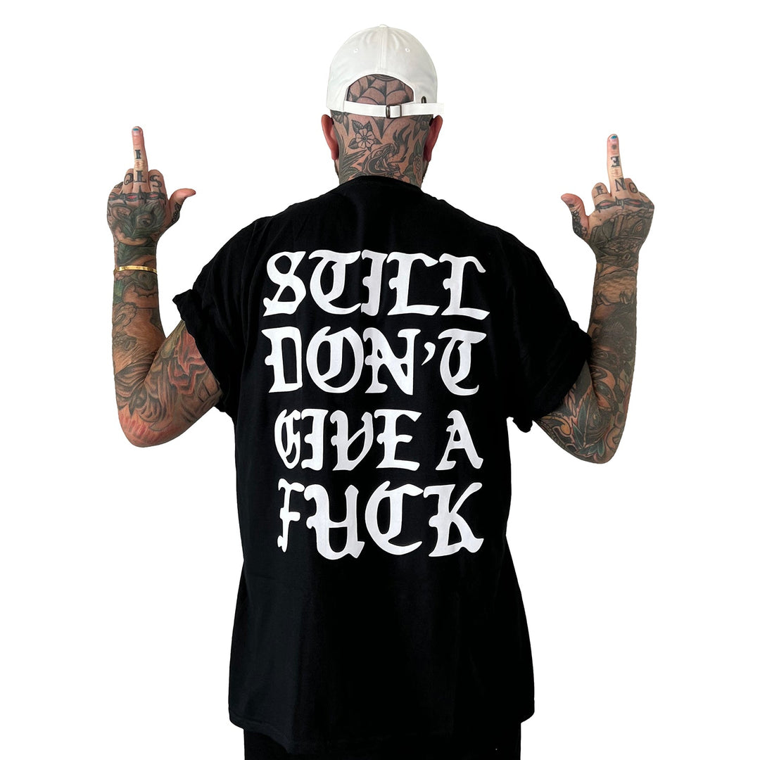 A tattooed individual in a white cap faces away, raising both middle fingers. Their black Stay Sick Clothing tee features bold white text: STILL DONT GIVE A FUCK.
