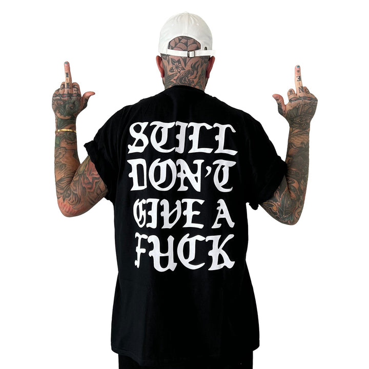 A tattooed individual in a white cap faces away, raising both middle fingers. Their black Stay Sick Clothing tee features bold white text: STILL DONT GIVE A FUCK.