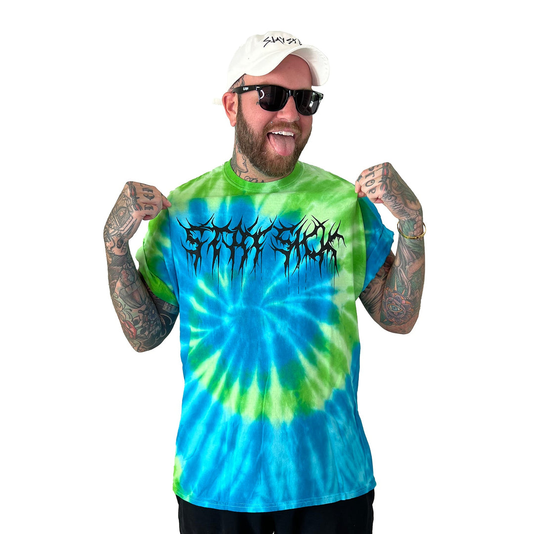 A smiling man with tattoos, sunglasses, and a white hat points at his blue-green Tie Dye T-Shirt with a bold Black Metal graphic from Stay Sick Clothing. The background is plain white.