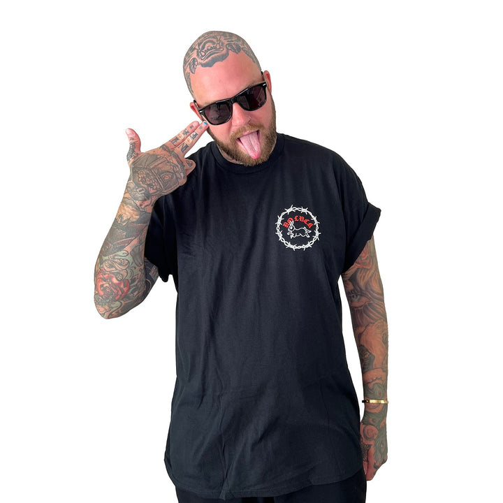 A tattooed, shaved-head man in sunglasses sticks out his tongue and makes a finger gun gesture to his head, wearing a black Stay Sick Clothing T-shirt with a white and red No Luck logo, standing against a white background.