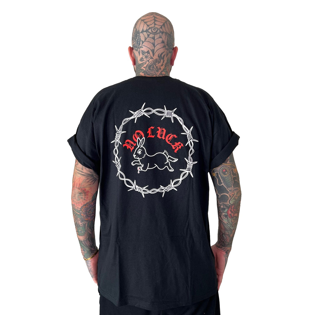 A tattooed individual faces away, wearing a Stay Sick Clothing Black T-Shirt. The shirt’s back features a white barbed wire circle, rabbit illustration, and bold red NO LUCK text, all set against a plain white background that highlights the striking design.