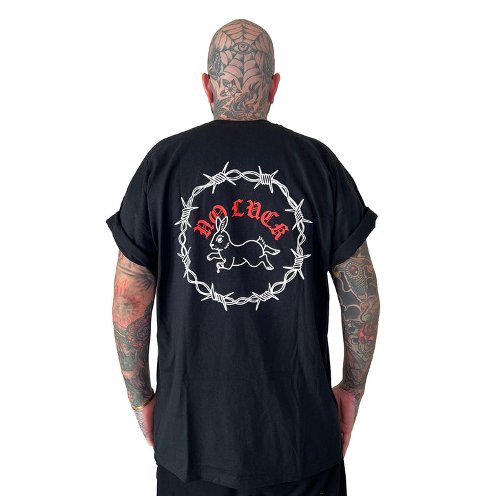 A tattooed individual faces away, wearing a Stay Sick Clothing Black T-Shirt. The shirt’s back features a white barbed wire circle, rabbit illustration, and bold red NO LUCK text, all set against a plain white background that highlights the striking design.