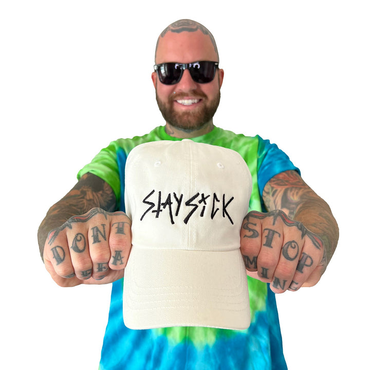 A smiling man in sunglasses and a tie-dye shirt holds a Stay Sick Clothing White Dad Hat with the SLAYSICK Scratch Logo. His tattooed fists, reading “DONT STOP” and more, are visible in the foreground.