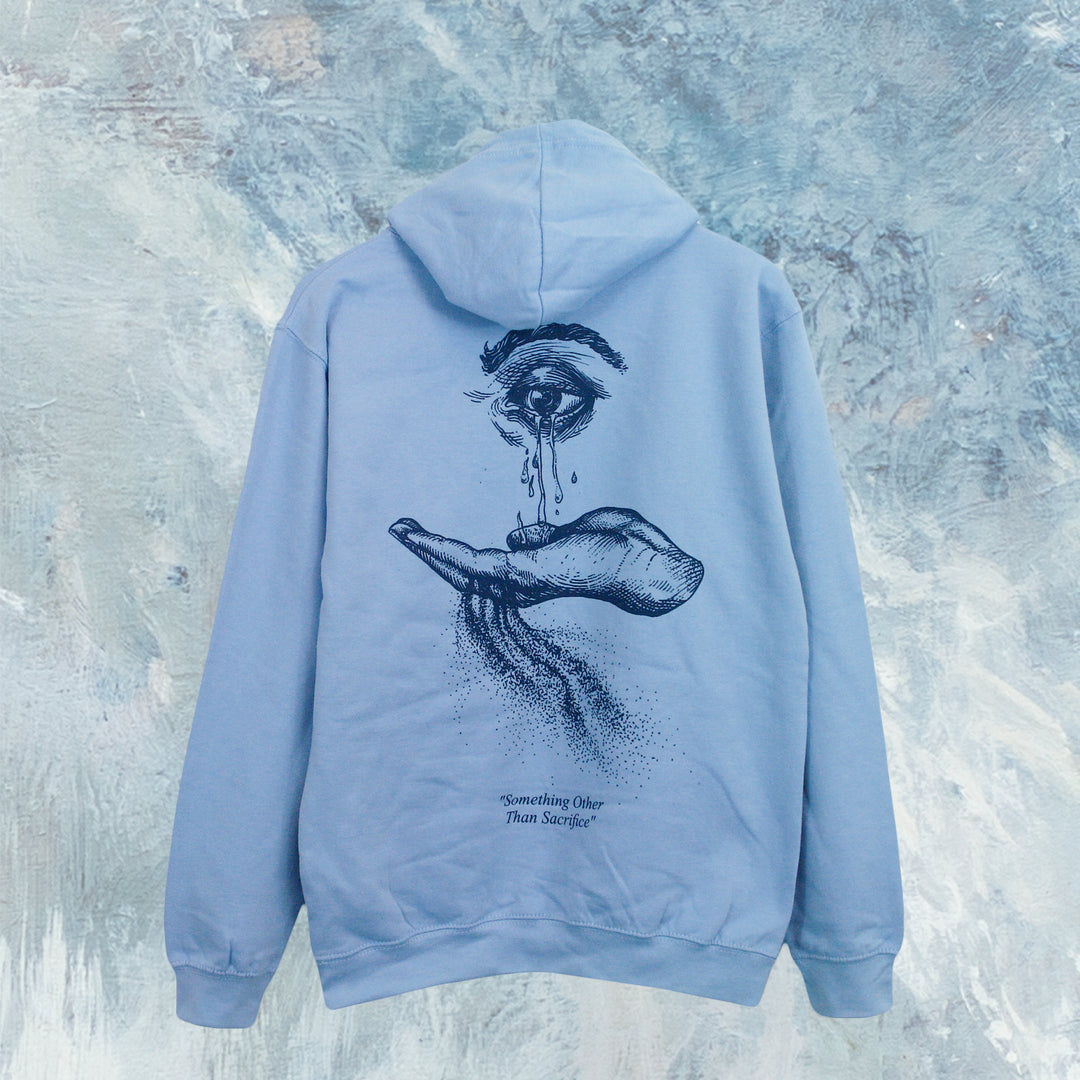 The Cryptodira light blue pullover hoodie features soft ringspun cotton, a black line art design of a large eye above an open hand with flowing tears on the back, and "Something Other Than Sacrifice" text below.