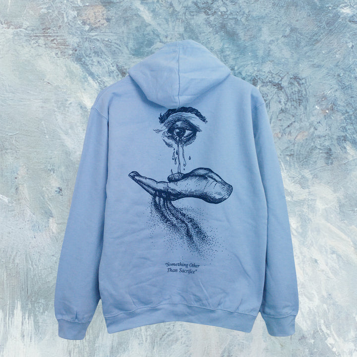 The Cryptodira light blue pullover hoodie features soft ringspun cotton, a black line art design of a large eye above an open hand with flowing tears on the back, and "Something Other Than Sacrifice" text below.