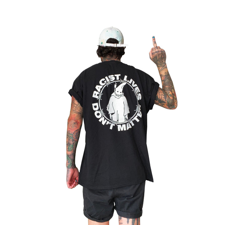 A tattooed individual wearing a black Stay Sick Clothing T-shirt that reads Racist Lives Don’t Matter with a graphic stands facing away, raising their middle finger and sporting a white cap and black shorts—a bold anti-racism statement.