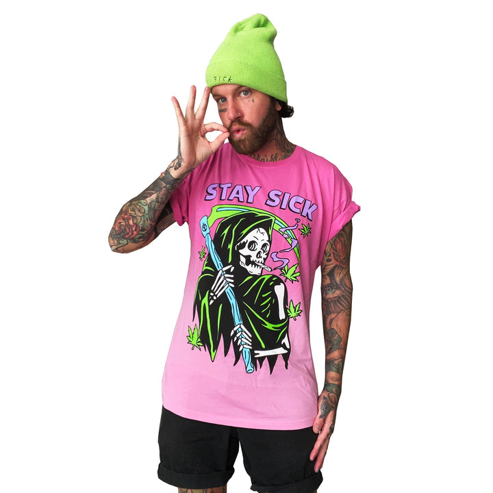 A tattooed person in a neon green beanie and a pink ombre Stay Sick Clothing t-shirt featuring a cartoon Grim Reefer making an OK sign stands against a white background.
