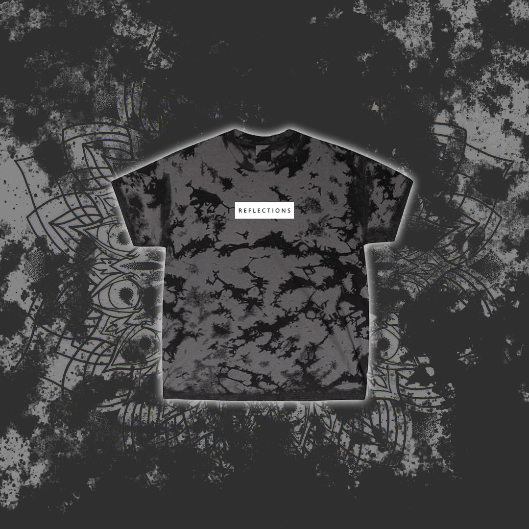 A charcoal black Reflections T-shirt with a white chest label and tie-dye pattern, displayed on a grunge-style gray and black abstract background with intricate line art.