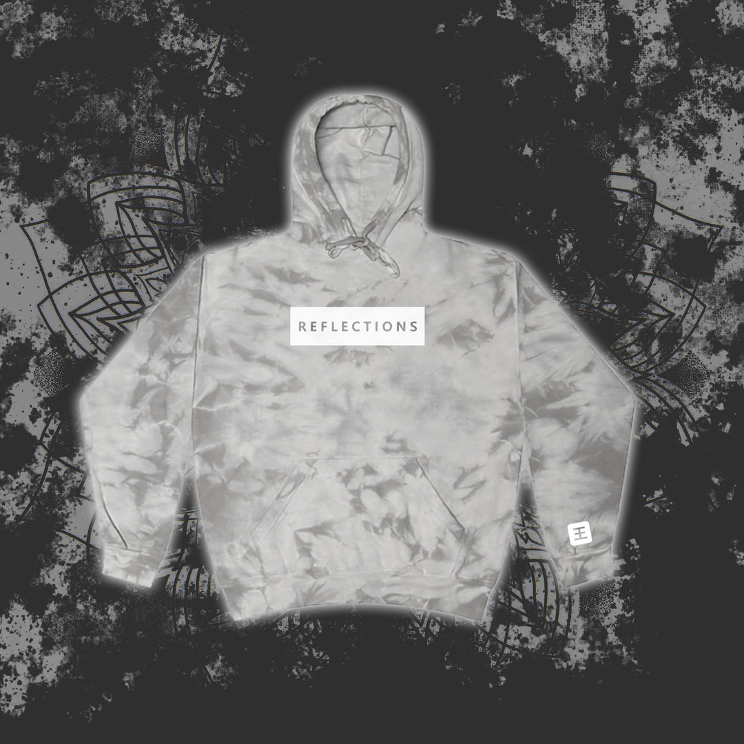 A gray Reflections tie-dye pullover hoodie with a white chest patch reading "REFLECTIONS" in black is showcased against a dark, abstract patterned background.