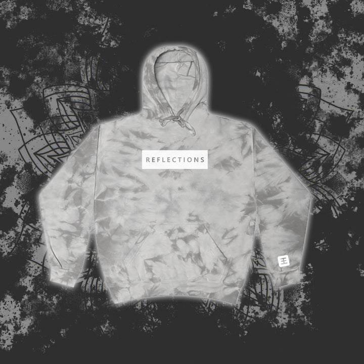 A gray Reflections tie-dye pullover hoodie with a white chest patch reading "REFLECTIONS" in black is showcased against a dark, abstract patterned background.