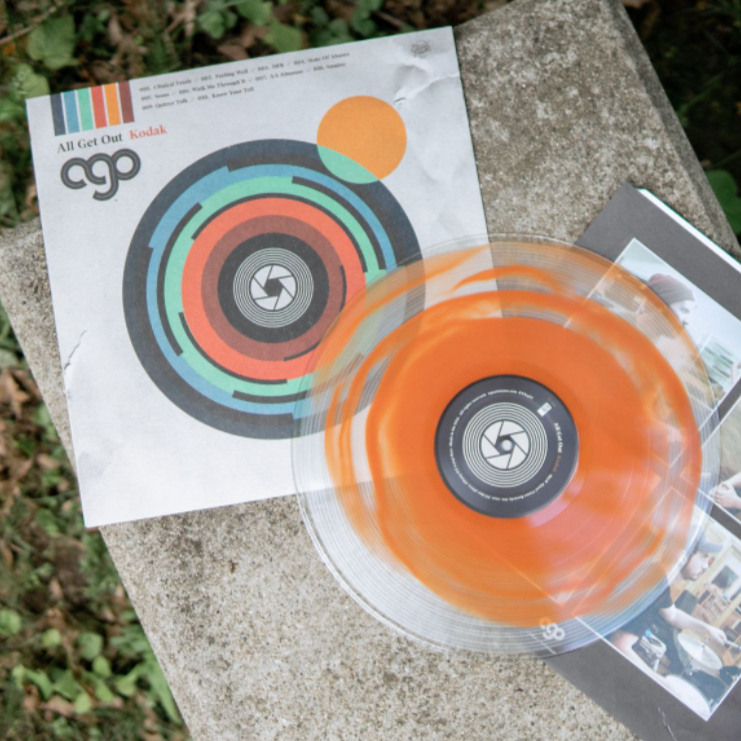 An orange and black swirl vinyl LP by All Get Out spins on a stone surface beside its vibrant geometric album cover, with greenery visible in the background.