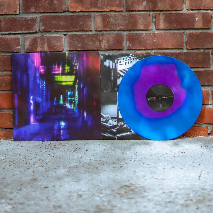 A blue and purple marbled Opaque Neon Magenta Vinyl of William Ryan Key’s Everything Except Desire, partially out of its sleeve with neon city art, stands against a red brick wall.