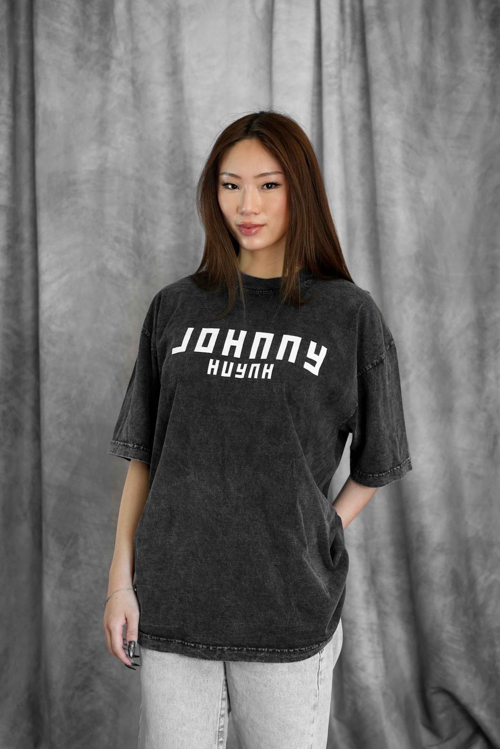 A woman with straight brown hair models a loose, unisex Johnny Huynh mineral wash T-shirt with white lettering and light jeans while standing before a gray fabric backdrop.