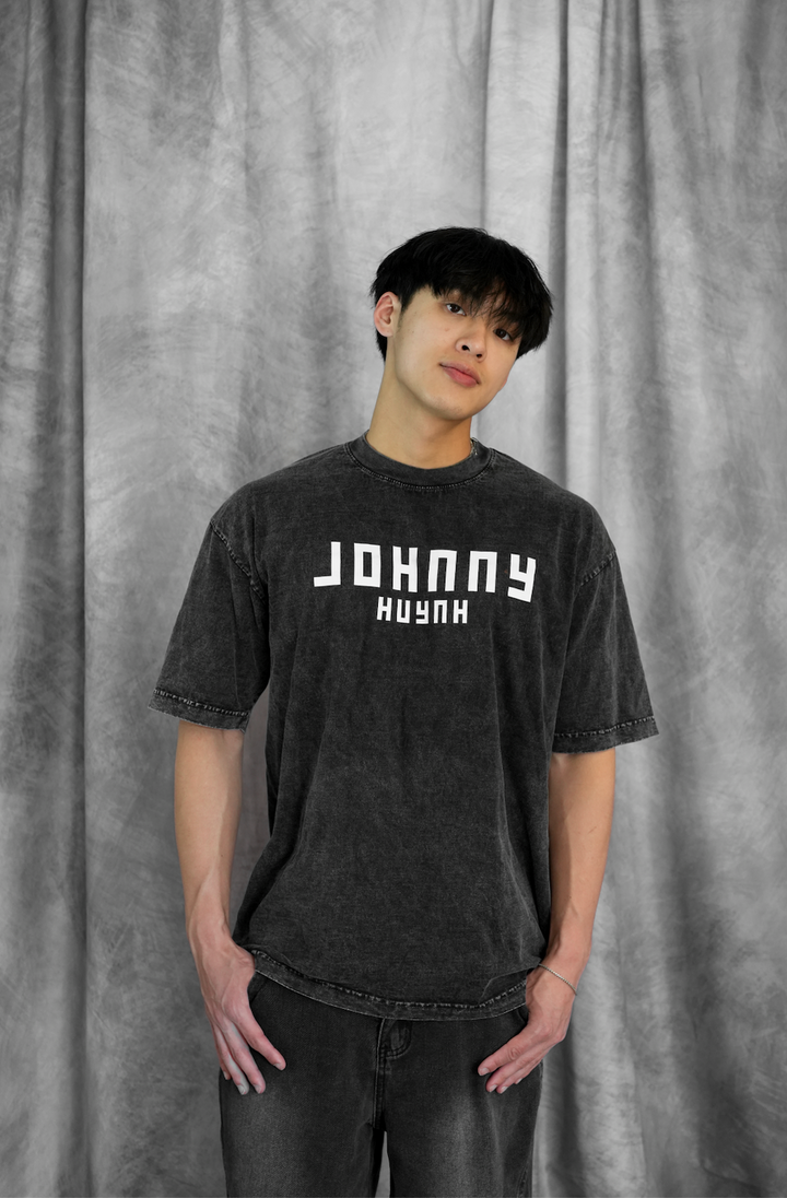 A young man stands before a gray backdrop, wearing a faded black unisex mineral wash t-shirt with bold white Johnny Huynh branding and dark jeans, hands in pockets, looking at the camera.