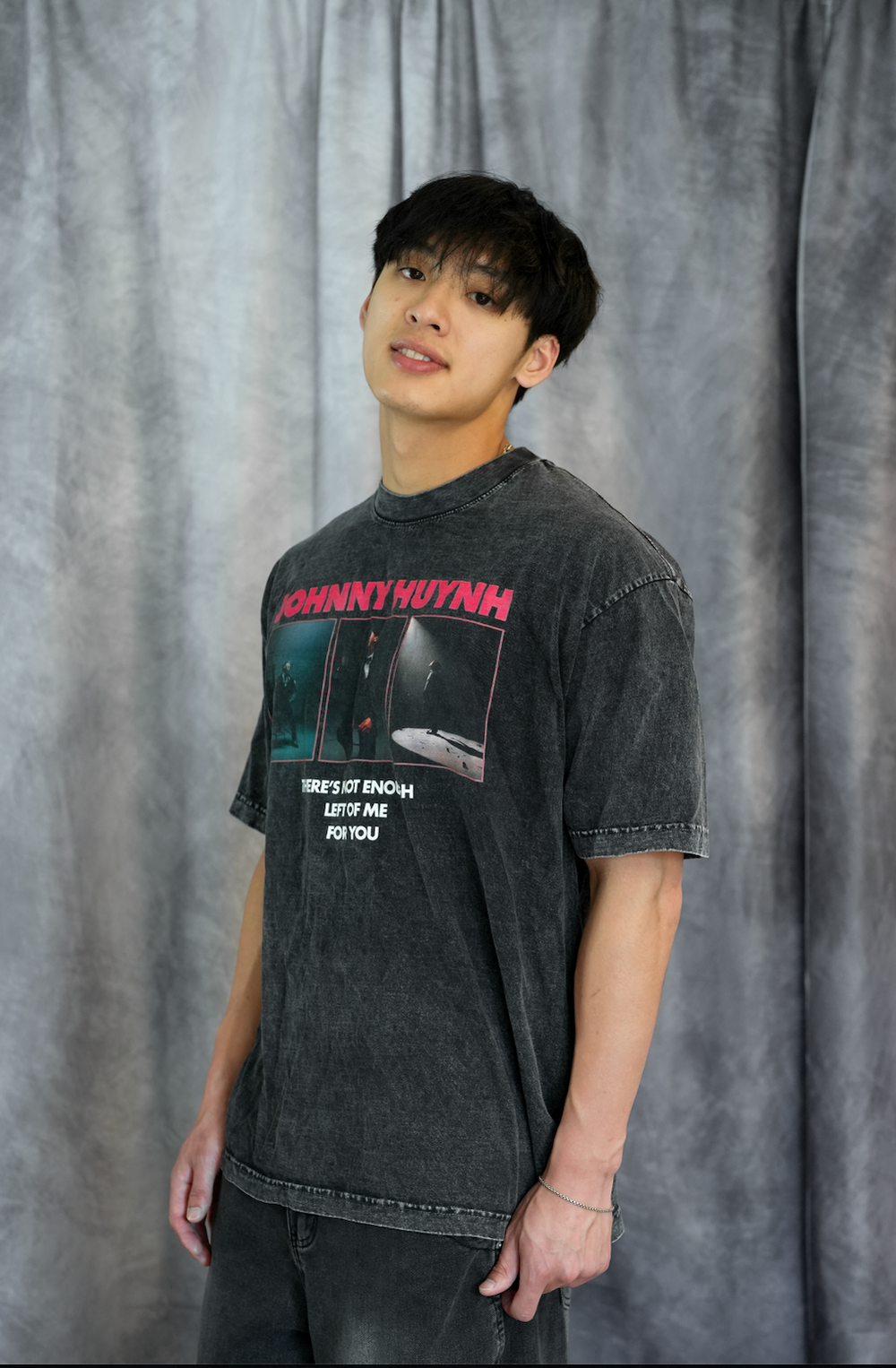 A young man with dark hair stands before gray curtains, wearing Johnny Huynh’s faded black mineral wash t-shirt and jeans. He faces slightly to the side, showcasing the brand’s unisex relaxed fit style with a calm expression.