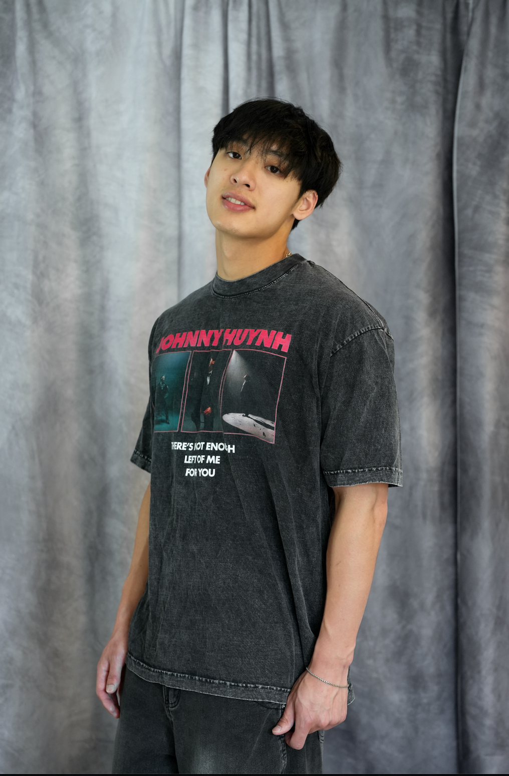 A young man with dark hair stands before gray curtains, wearing Johnny Huynh’s faded black mineral wash t-shirt and jeans. He faces slightly to the side, showcasing the brand’s unisex relaxed fit style with a calm expression.