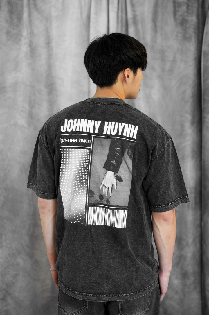 A person with short black hair faces away, wearing a black Johnny Huynh unisex garment-dyed cotton t-shirt. The back graphic reads "JOHNNY HUYNH jah-nee hwin" above a gloved hand photo and barcode design. Gray curtain backdrop.
