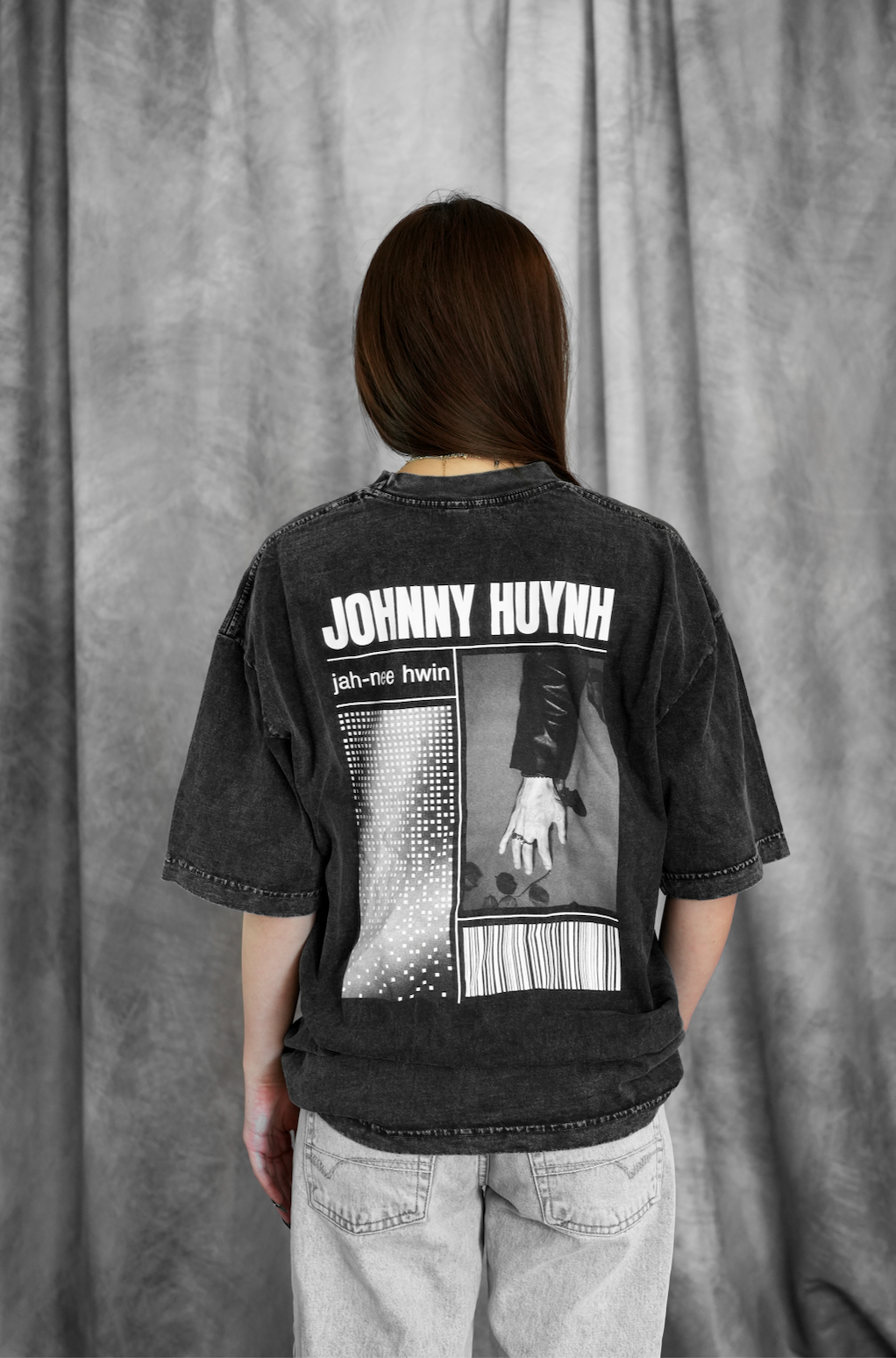 A person with long brown hair stands facing a gray curtain, wearing a Johnny Huynh black unisex graphic T-shirt made from garment-dyed cotton, featuring text and designs on the back, paired with light gray jeans.