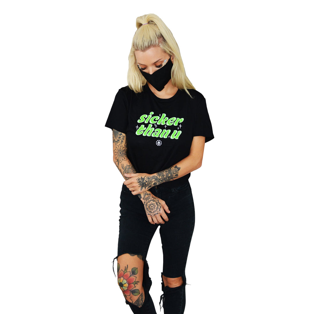 A woman with blonde hair in a high ponytail wears a Stay Sick Clothing black cropped T-shirt with “sicker than u” in neon green, ripped black jeans, and a mask. Tattoos on her arms and leg add to the edgy vibe—perfect for your Black Girl’s T-Shirt collection.