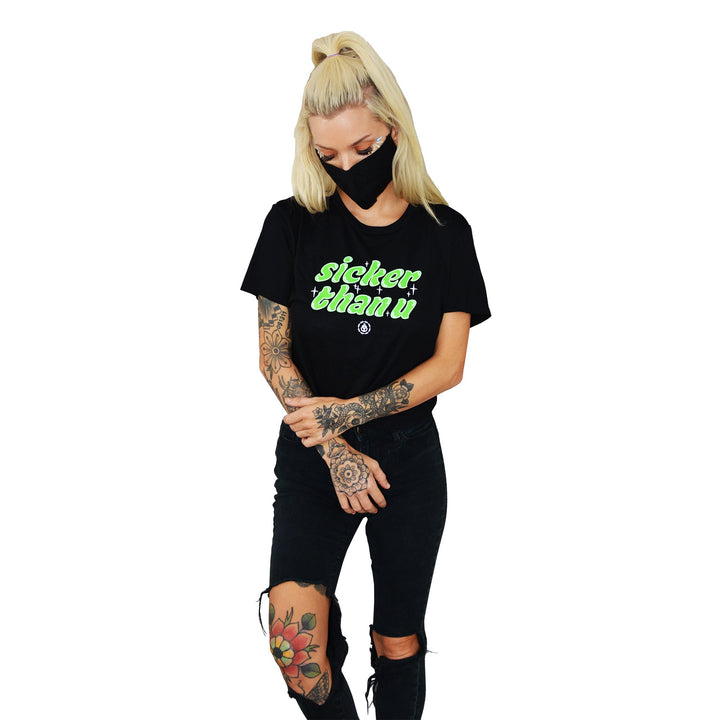 A woman with blonde hair in a high ponytail wears a Stay Sick Clothing black cropped T-shirt with “sicker than u” in neon green, ripped black jeans, and a mask. Tattoos on her arms and leg add to the edgy vibe—perfect for your Black Girl’s T-Shirt collection.