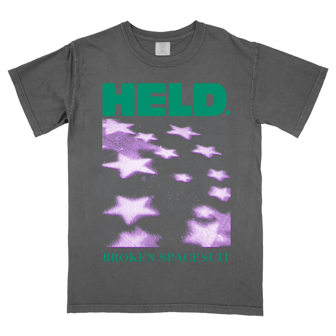 Gray Comfort Colors t-shirt with green text reading HELD. at the top and BROKEN SPACESUIT at the bottom. The center features blurred, purple star shapes scattered diagonally—a unique choice for fans of printed shirts.