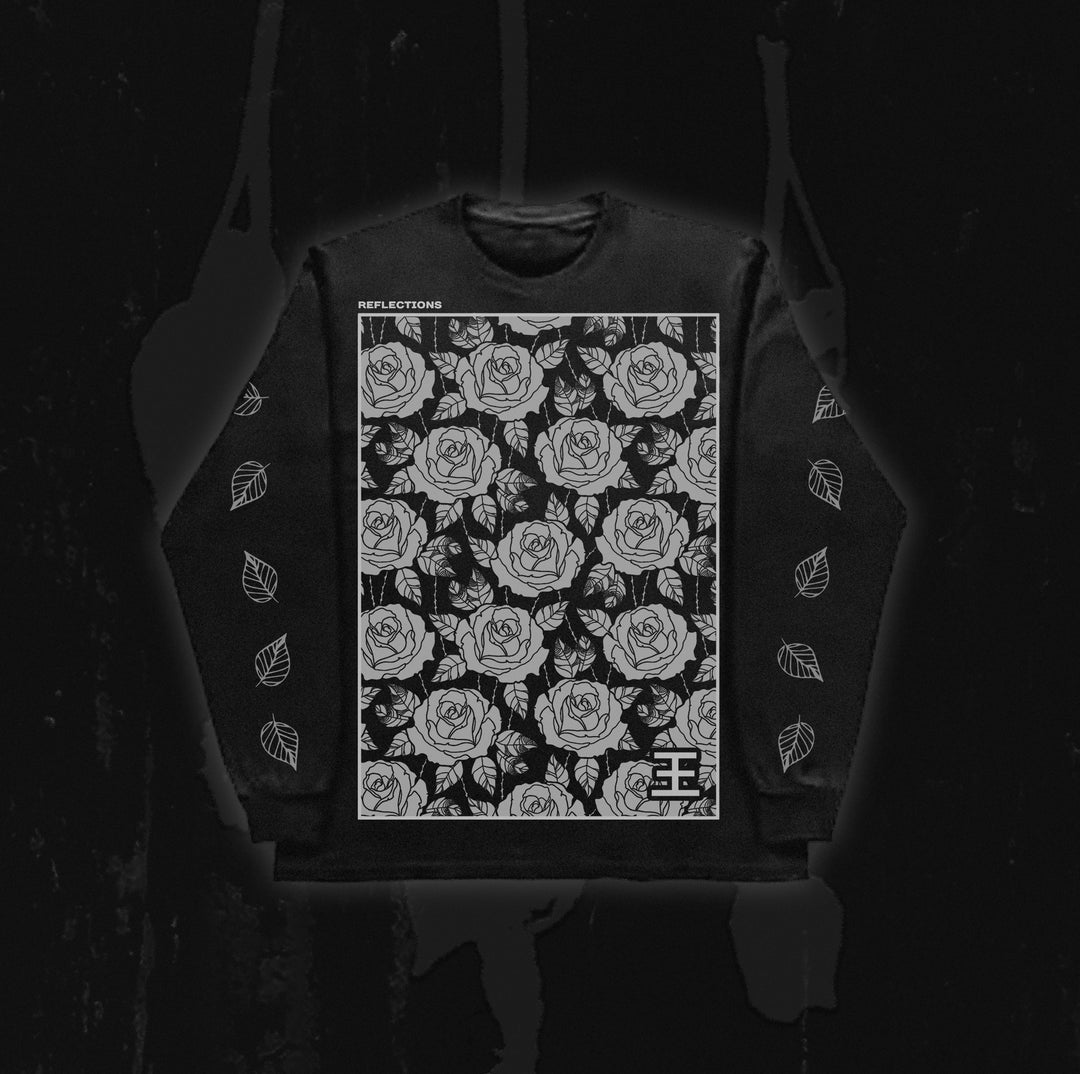 A black Reflections sweatshirt showcases a large white rose pattern on the front with “REFLECTIONS” text across the top, inspired by the ethereal style of Crystal Eyes. White leaf designs adorn both sleeves against a dark background.