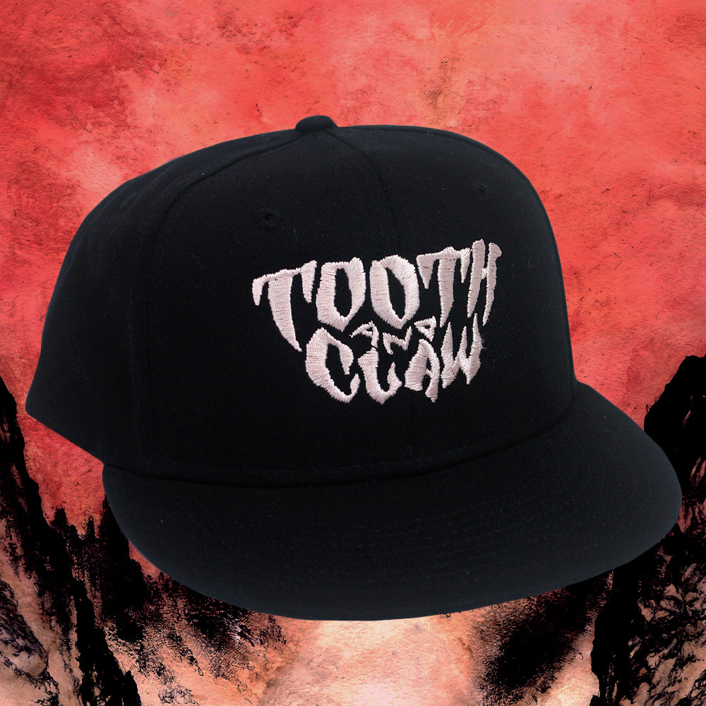 A black Tooth And Claw Flat Bill Cap snapback with bold white TOOTH AND CLAW text sits on a textured red and black background, made from durable Cotton Twill in a structured 6-panel design.
