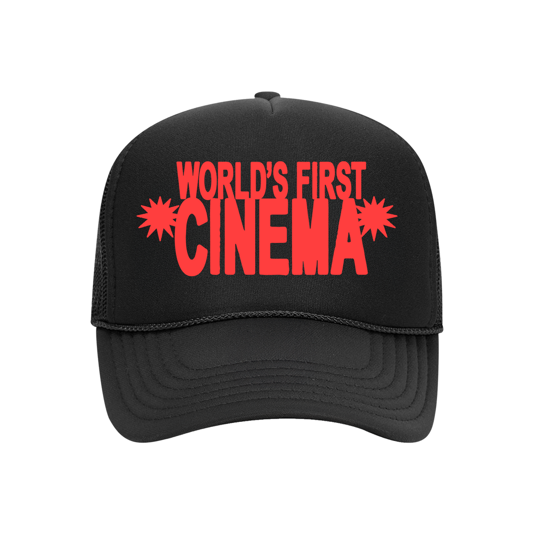 A black trucker hat from World's First Cinema featuring bold red stacked text "WORLDS FIRST CINEMA" and starburst designs beside "CINEMA," all on breathable black mesh.