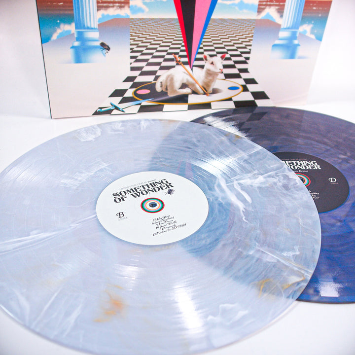 Two marbled vinyl records—a Double LP in light and dark blue from World's First Cinema—are displayed on a white surface. The Something of Wonder album cover, featuring a white cat and abstract shapes, is in the background.