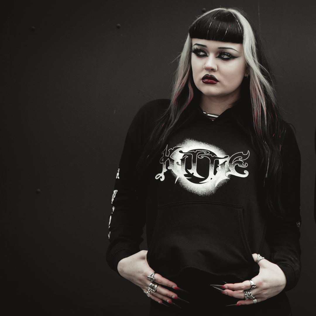 A person with dramatic black and white hair, dark eye makeup, and black lipstick wears a Kittie women’s cropped hoodie made from AWDis ringspun cotton with a white gothic design, standing seriously against a dark background.