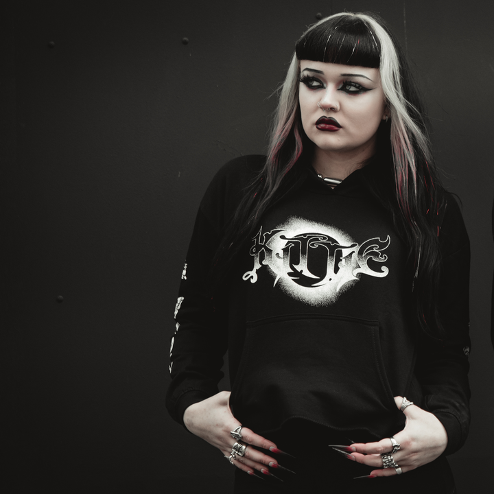 A person with dramatic black and white hair, dark eye makeup, and black lipstick wears a Kittie women’s cropped hoodie made from AWDis ringspun cotton with a white gothic design, standing seriously against a dark background.