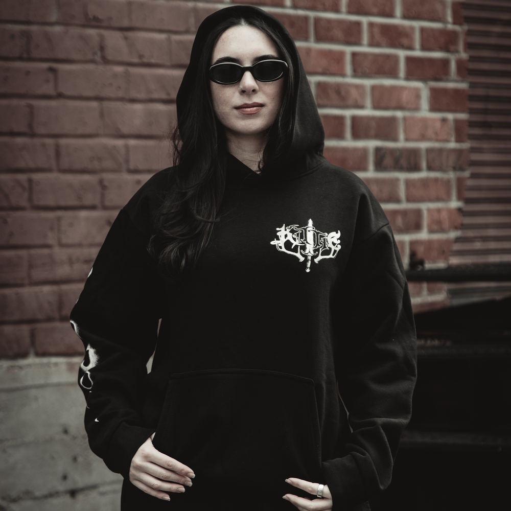 A woman with long dark hair and dark sunglasses stands confidently before a red brick wall, wearing an oversized fit Kittie Element Hoodie featuring white designs.