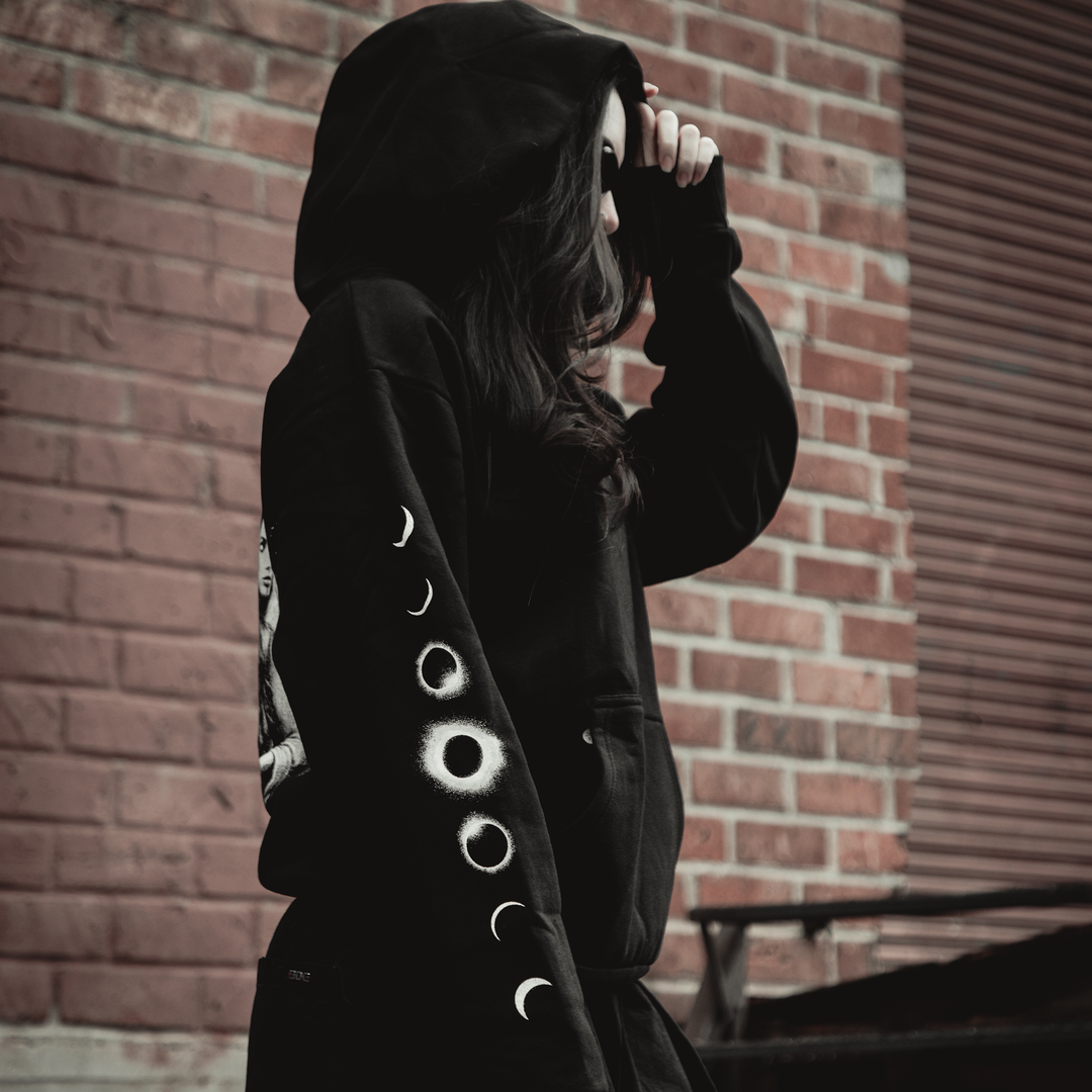A person wearing a black Kittie Element Hoodie with moon phases on the sleeve stands by a brick wall, holding the oversized hood over their head and looking away from the camera.