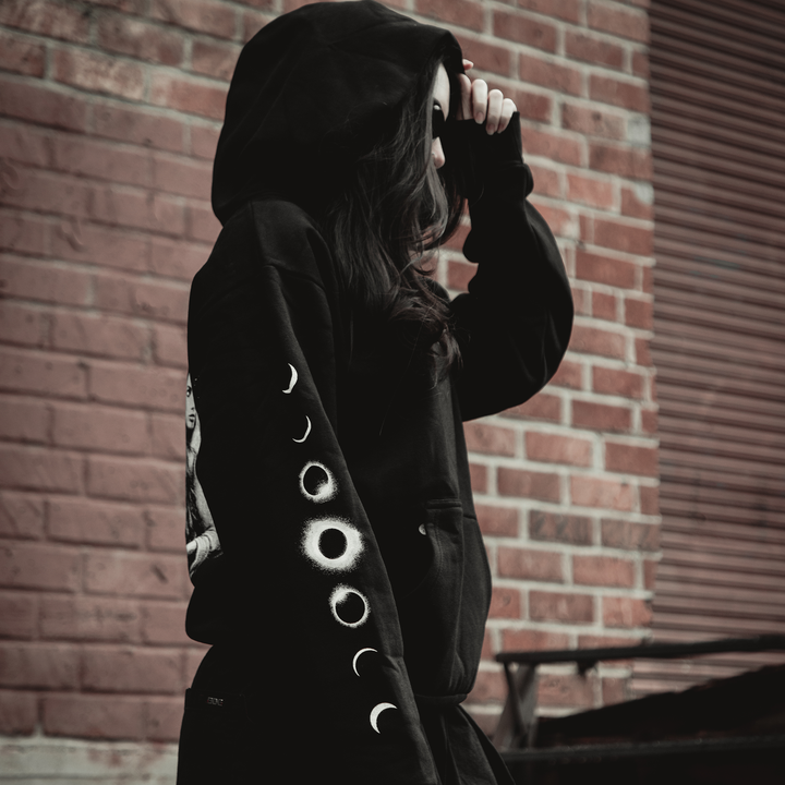 A person wearing a black Kittie Element Hoodie with moon phases on the sleeve stands by a brick wall, holding the oversized hood over their head and looking away from the camera.