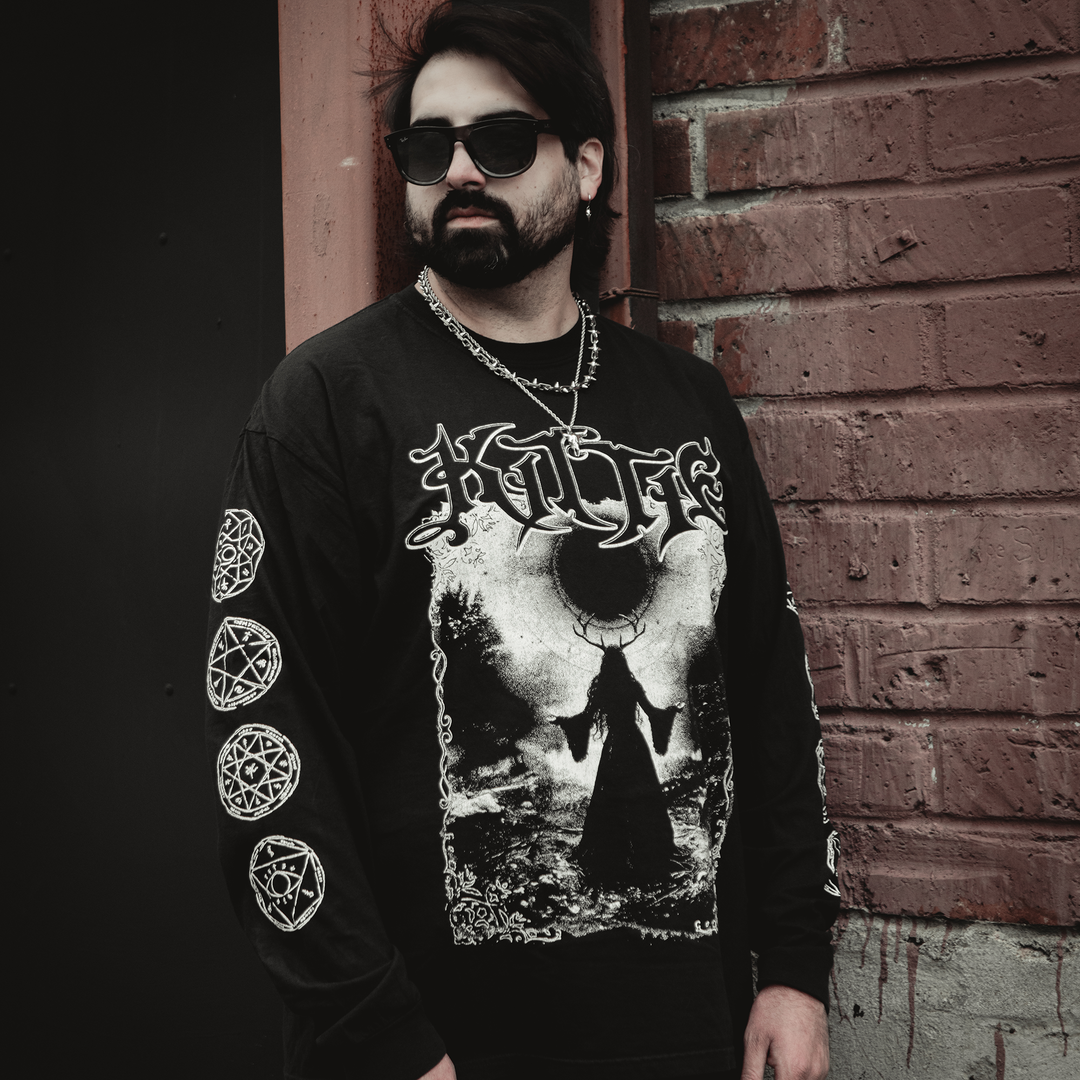 A bearded person with long dark hair and sunglasses stands by a brick wall, wearing a Kittie Comfort Colors black long sleeve shirt made from garment-dyed, ring spun cotton featuring occult-themed graphics and a chain necklace.