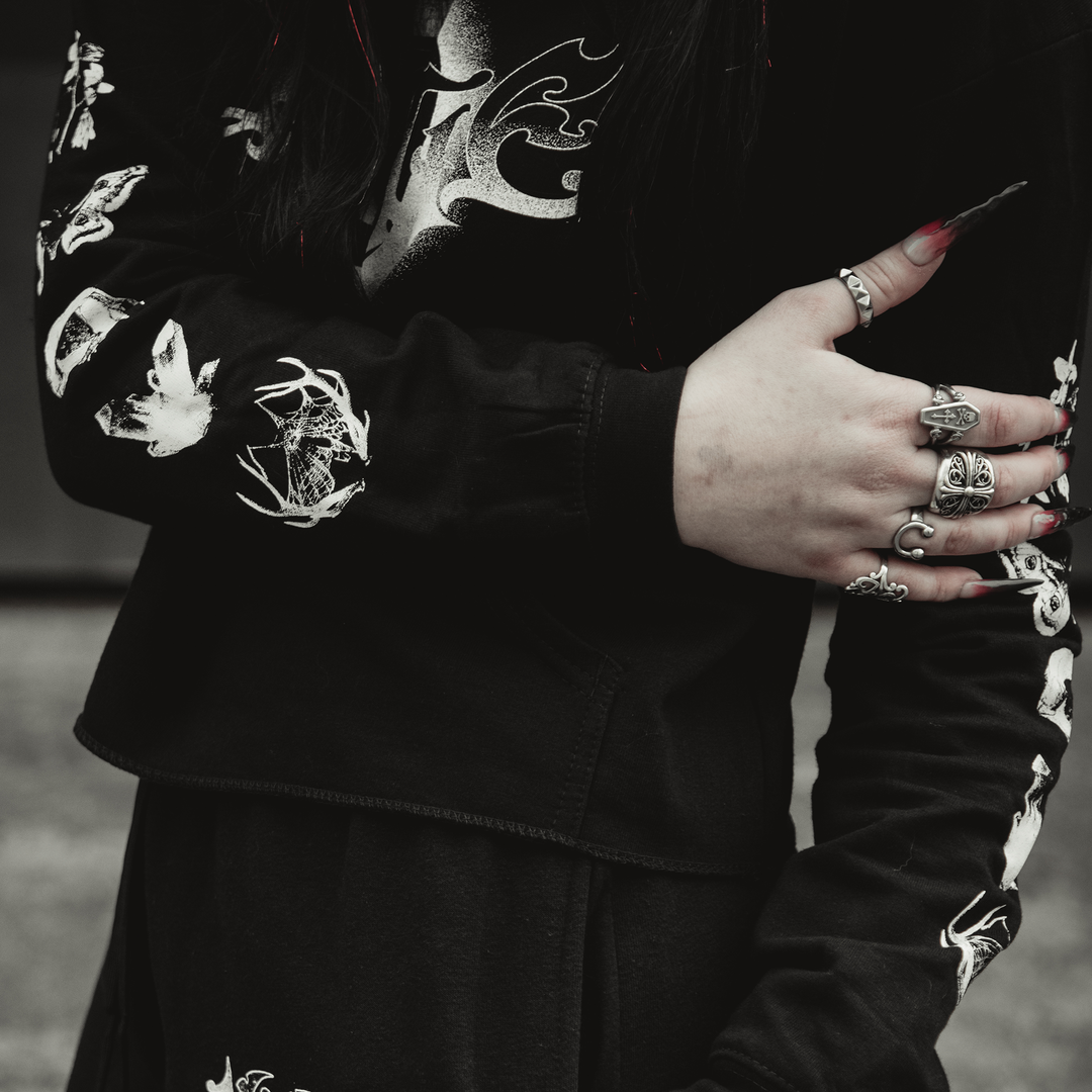 A person in a Kittie black long-sleeve ringspun cotton shirt with white graphics crosses one arm over their body, showing long painted nails and multiple silver rings. The background is blurred.