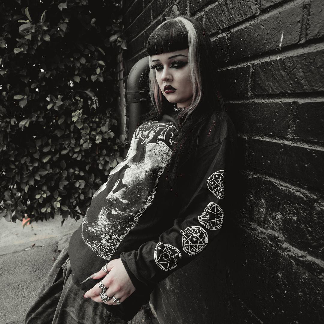 A person with black and white hair, dark makeup, and gothic clothing leans against a brick wall. They wear Kittie garment-dyed Comfort Colors long sleeves with occult-style graphics. A bush is in the background.