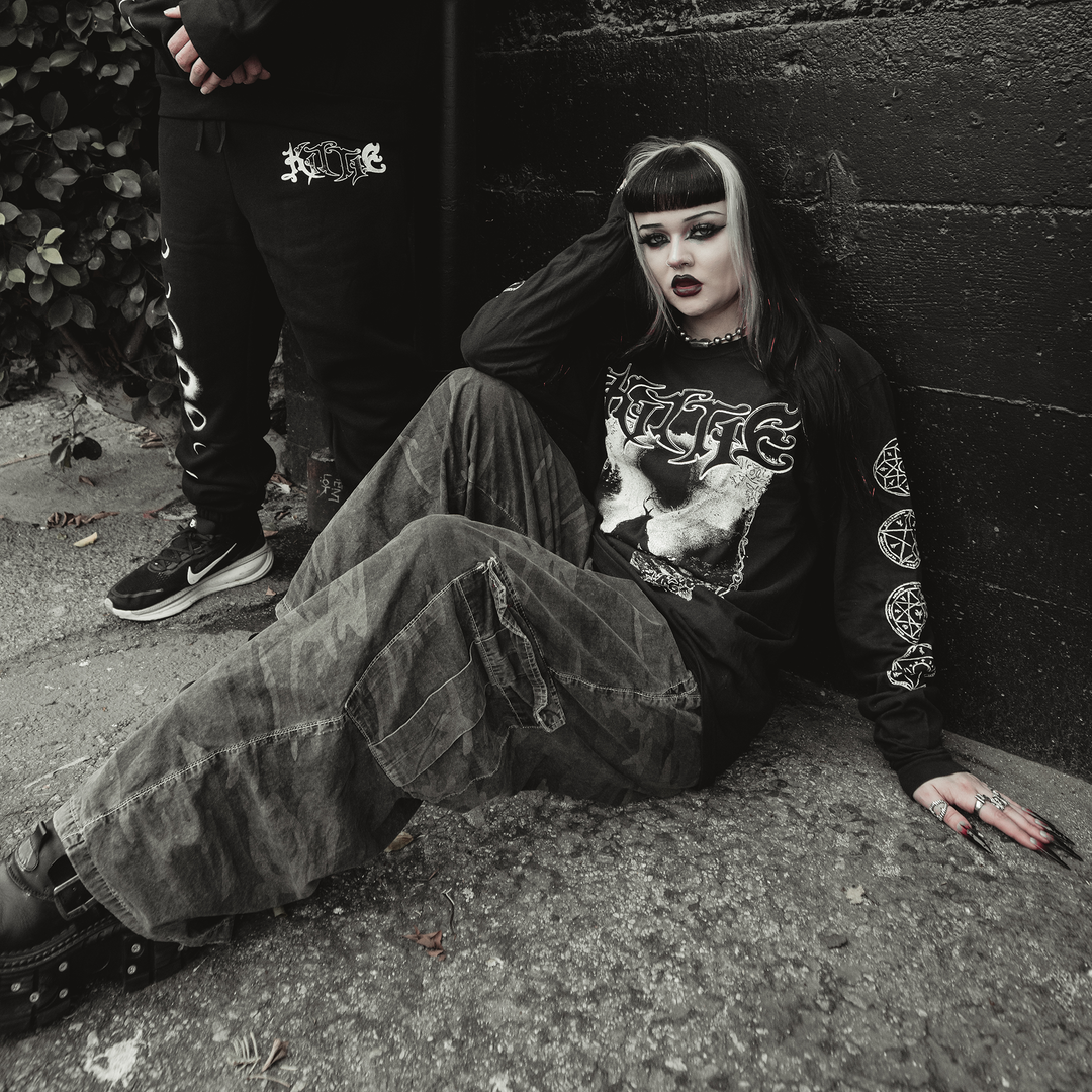 A person with black and white hair, dark makeup, and gothic clothing sits on the ground against a black wall. Nearby, someone wearing a garment-dyed Kittie Comfort Colors long sleeve stands partially out of frame.