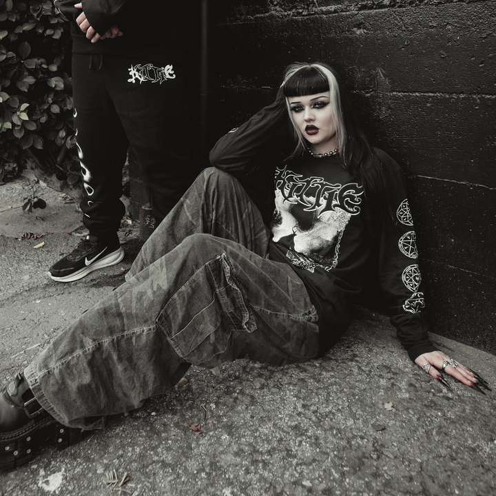 A person with black and white hair, dark makeup, and gothic clothing sits on the ground against a black wall. Nearby, someone wearing a garment-dyed Kittie Comfort Colors long sleeve stands partially out of frame.