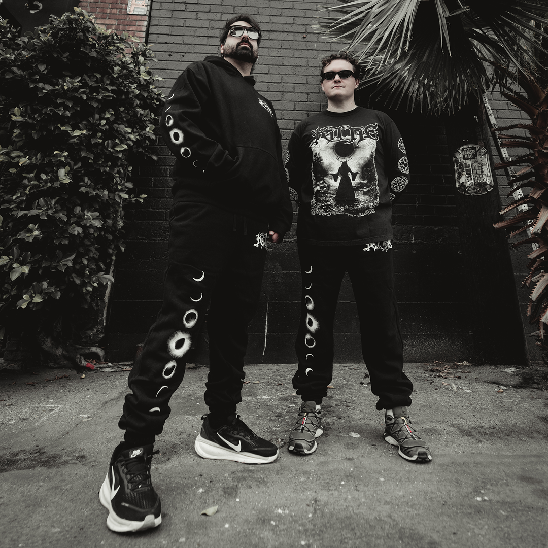 Two people stand confidently outdoors against a dark wall, both dressed in Kittie’s black moon-phase streetwear made from recycled polyester with bold graphic prints. Dark sunglasses and chunky sneakers complete their look, framed by palm leaves and brickwork.